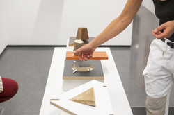 Rundgang Kunst Holz Auf einem niedrigen, weißen Block sind vier Plastiken aus Holz ausgestellt. Links im Bild ist das Knie einer hockenden Person zu sehen. Rechts sieht man Ausschnitte einer Person, die über den Plastiken gestikuliert.