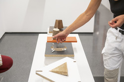 Auf einem niedrigen, weißen Block sind vier Plastiken aus Holz ausgestellt. Links im Bild ist das Knie einer hockenden Person zu sehen. Rechts sieht man Ausschnitte einer Person, die über den Plastiken gestikuliert.