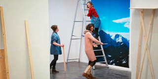 Drei Frauen bringen eine großformatige Fotografie an einer Wand an, im Vordergrund stehen Werkzeuge und Wandfarben