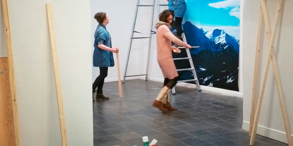 Drei Frauen bringen eine großformatige Fotografie an einer Wand an, im Vordergrund stehen Werkzeuge und Wandfarben
