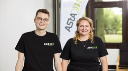 Ein Mann und eine Frau, die beide ein schwarzes T-Shirt mit einem Logo des AStA der TU Dortmund tragen, sitzen auf einem Tisch. Hinter Ihnen ist ein Aufsteller, auf dem AStA geschrieben steht.