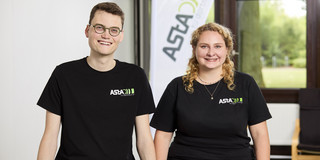 Ein Mann und eine Frau, die beide ein schwarzes T-Shirt mit einem Logo des AStA der TU Dortmund tragen, sitzen auf einem Tisch. Hinter Ihnen ist ein Aufsteller, auf dem AStA geschrieben steht.