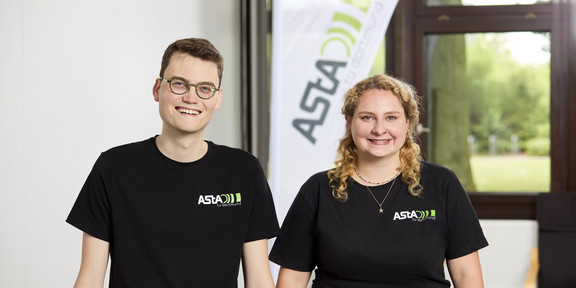 Ein Mann und eine Frau, die beide ein schwarzes T-Shirt mit einem Logo des AStA der TU Dortmund tragen, sitzen auf einem Tisch. Hinter Ihnen ist ein Aufsteller, auf dem AStA geschrieben steht.