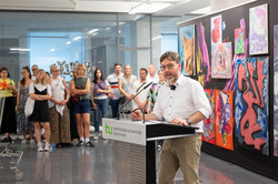 Im Zentrum des Bildes steht Dr. Niklas Gliesmann an einem Pult. Hinter ihm befindet sich eine schwarze Wand mit Kunstwerken von Studierenden sowie Kunstinteressierten.