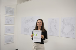 Eine Frau steht vor einer weißen Wand an der mehrere Bilder einer Kunstausstellung hängen und hält eine Urkunde hoch.