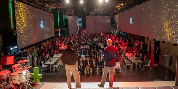 Photo: Two people stand on a stage and address a large audience in an event hall.