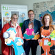 Prof. Prof. Barbara Welzel, Prof. Thomas Goll und Svenja Sayk stehen vor einem großen Aufsteller der KinderUni.