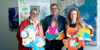 Prof. Prof. Barbara Welzel, Prof. Thomas Goll und Svenja Sayk stehen vor einem großen Aufsteller der KinderUni.