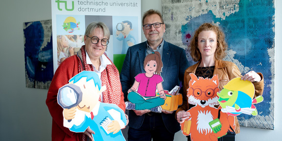 Prof. Prof. Barbara Welzel, Prof. Thomas Goll und Svenja Sayk stehen vor einem großen Aufsteller der KinderUni.