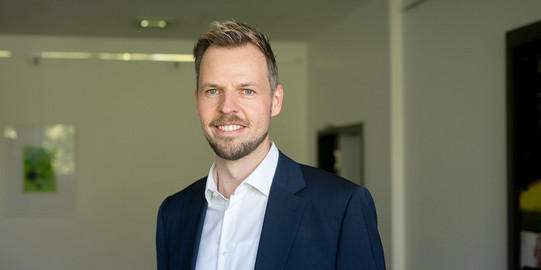 Zu sehen ist ein Porträtfoto von Prof. Peitz in einem Raum mit weißen Wänden im Hintergrund.