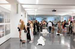 Das Bild zeigt einen großen Saal mit vielen Menschen. Auf der linken Seite befinden sich Bilder auf einer weißen Wand. Im Zentrum des Bildes sind weiße Kunstinstallationen auf dem Boden liegend zu sehen.