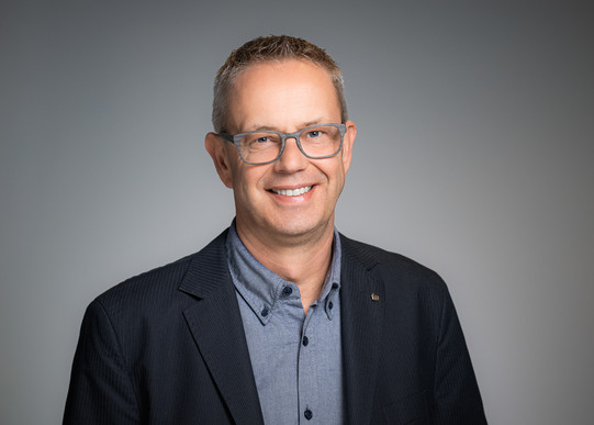 Portrait photo of Chancellor Markus Neuhaus: Markus Neuhaus wears a dark jacket and a patterned shirt and stands in front of a gray background. He smiles into the camera.