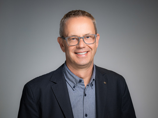 Portrait photo of Chancellor Markus Neuhaus: Markus Neuhaus wears a dark jacket and a patterned shirt and stands in front of a gray background. He smiles into the camera.