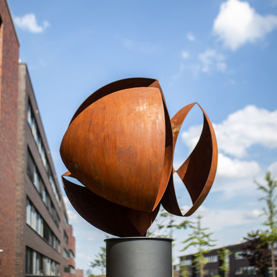Skulptur Eine rundliche, bronzefarbene Skulptur steht auf einem Sockel. Im Hintergrund blauer Himmel, Bäume und Teil eines Gebäudes.