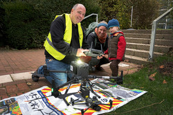 Im Vordergrund des Bildes ist eine Drohne auf einem plakatartigen Untergrund zu sehen. Dahinter befinden sich zwei erwachsene Personen und ein Kind.