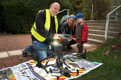 Im Vordergrund des Bildes ist eine Drohne auf einem plakatartigen Untergrund zu sehen. Dahinter befinden sich zwei erwachsene Personen und ein Kind.