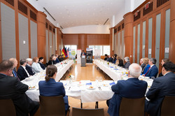 20 Jahre New York Büro - Auftaktrede Auf dem Bild ist ein großer Saal mit einem u-förmig angeordneten Tisch zu sehen, an dem viele Personen in Anzügen sitzen.