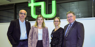 Group photo: Four people stand in a room in front of a large, brightly lit green TU logo.
