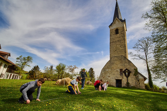 Auf dem Foto sieht man Kinder auf einer Wiese vor der Kirche mit Zollstöcken etwas ausmessen.