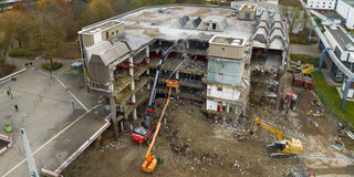 Eine Luftaufnahme von einer Baustelle an der Mensabrücke. Das Gemäuer der alten Zentralbibliothek wird von einem Spezialbagger abgetragen während von einem Kran aus jemand das Abbruchgut bewässert.