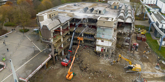 Eine Luftaufnahme von einer Baustelle an der Mensabrücke. Das Gemäuer der alten Zentralbibliothek wird von einem Spezialbagger abgetragen während von einem Kran aus jemand das Abbruchgut bewässert.