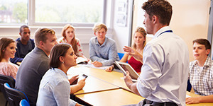 Fellowship Dozent ist an einen Tisch gelehnt und spricht zu daran sitzenden Studierenden.