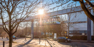 Das Bild zeigt die H-Bahn der TU Dortmund. Direkt darüber strahlt die Sonne. Im Vordergrund befindet sich ein rot gepflasteter Weg, ein bisschen Grünfläche sowie kahle Bäume. Dahinter folgt die Emil-Figge-Straße. Hinter der H-Bahn ist ein weißes Gebäude erkennbar.