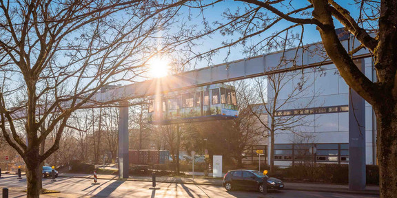Das Bild zeigt die H-Bahn der TU Dortmund. Direkt darüber strahlt die Sonne. Im Vordergrund befindet sich ein rot gepflasteter Weg, ein bisschen Grünfläche sowie kahle Bäume. Dahinter folgt die Emil-Figge-Straße. Hinter der H-Bahn ist ein weißes Gebäude erkennbar.