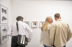 Rundgang Kunst Fotowürfel und Kleinportraits Ein Mann steht links und beugt sich vor, um fünf Kleinportraits zu betrachten. Rechts stehen eine Frau und ein Mann und unterhalten sich über die Bilder.