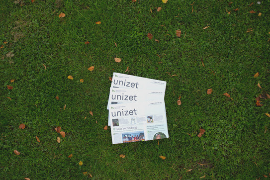 Three issues of unizet from August 2023 lie fanned out on top of each other in the grass.