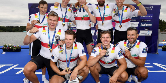 Deutschlandachter Gruppenfoto von neun Männern, die Goldmedaillen in die Kamera halten