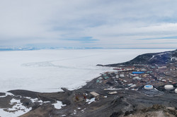 Numerous smaller buildings and containers on brown earth in front of an ice shelf.