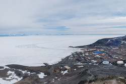 McMurdo Research Station Numerous smaller buildings and containers on brown earth in front of an ice shelf.