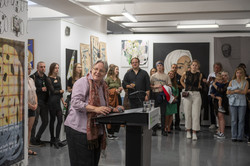 Rundgang Kunst Prof. Barbara Welzel Prof. Barbara Welzel steht an einem Rednerpult. Im Hintergrund stehen Menschen vor den ausgestellten Kunstwerken.