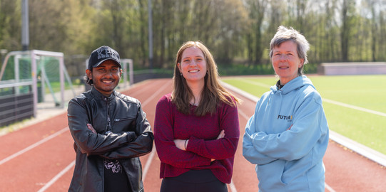 Gruppenfoto von Volunteers bei den FISU Games 2025: Das Foto zeigt drei Freiwillige mit verschränkten Armen auf einer Laufbahn. Von links nach rechts: Ein Student mit schwarzer Kappe und getönter Brille, eine Studentin mit braunen Haaren und rotem Pullover und eine Seniorenstudentin mit kurzen Haaren und hellblauer Jacke.