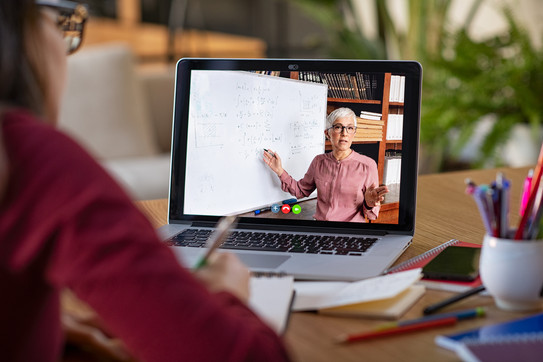 Mädchen sitzt an einem Schreibtisch und schaut auf einen Laptop, auf dem eine Frau in einem Video-Chat zu sehen ist.