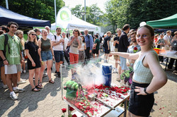 Rechts steht eine Studentin mit Brille und grün-weiß karriertem Crop-Top, die ihre linke Hand in die Hüfte stützt, mit der rechten Hand eine Rose in einen dampfenden, blauen Metallzylinder hält und in die Kamera lächelt. Vor ihr ist ein mit Plexiglas eingerahmter Tisch, auf dem ganz viele zerkleinerte, rote Rosenblütenblätter verstreut liegen. Ein kleiner Metallhammer mit Verzierung liegt dazwischen. Links hinter dem Tisch steht eine Gruppe junger Menschen, die zuschauen und lächeln.