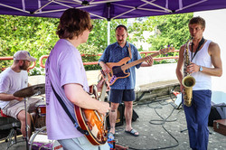 A band consisting of a drummer, bassist, guitarist and saxophonist playing live music under a pavilion.