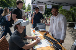 Ein junger Mann trägt eine VR-Brille und hält einen Controller. Die drei weiteren Männer, die am Stand stehen, sehen ihm zu und lächeln.