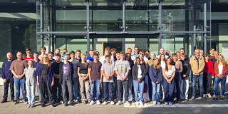 Gruppenfoto von jungen Auszubildenden und ihren Ausbilder*innen in der Sonne vor einem Universitätsgebäude mit Glasfront.