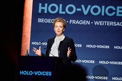 Foto: Eine Person spricht an einem Rednerpult vor einer blauen Wand mit dem Schriftzug „HOLO-VOICES“.