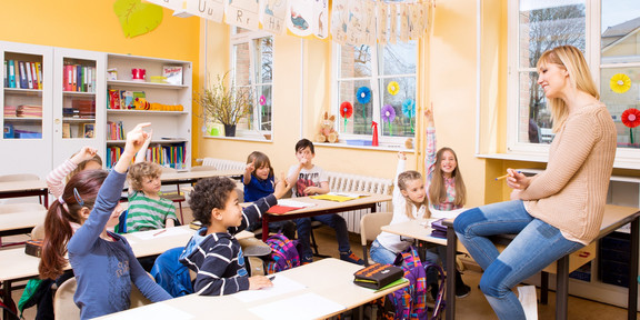 Foto: Blick ins Klassenzimmer - Kinder sitzen an Tischen, vorne sitzt die Lehrerin auf dem Pult.