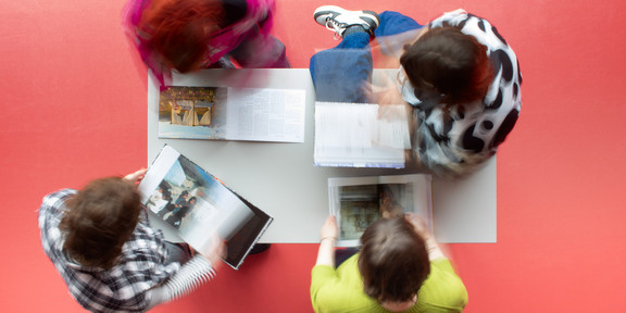 Von oben fotografiert: Vier Personen um einen Tisch herum stehend und sitzend, alle vier lesen Bücher