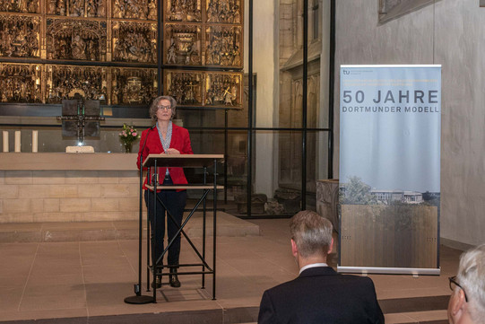 Das Bild zeigt die NRW-Bauministerin Ina Scharrenbach in der Stadtkirche St. Petri in Dortmund hinter einem Pult. Rechts von ihr steht ein Aufsteller mit "50 Jahre Bauwesen". Hinter ihr befindet sich der Altar der Kirche.