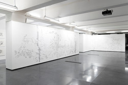Im Zentrum des Bildes steht eine weiße, große Ausstellungswand, die komplett bezeichnet ist.