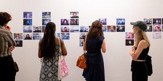 Vier Personen stehen vor einer weißen Wand, auf der einige Fotos angebracht sind