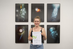Eine Frau steht vor einer weißen Wand an der mehrere Bilder einer Kunstausstellung hängen und hält eine Urkunde hoch.