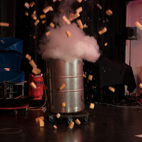 Eine große Tonne, aus der dichter Rauch strömt. Daneben steht ein Mann auf einer Bühne, der die Explosion beobachtet.