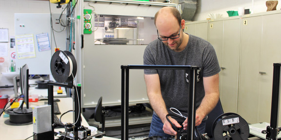 A man stands at a table in a workshop and operates a 3D printer.