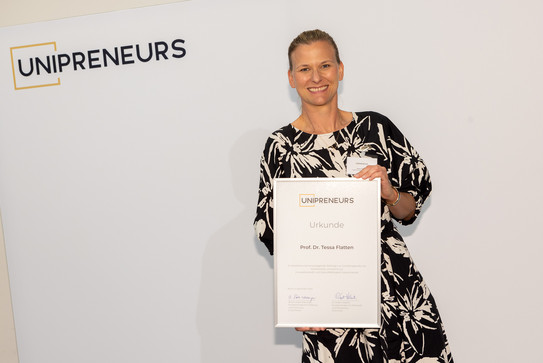 Unipreneurs Flatten Eine Frau, Prof. Tessa Flatten, hält ihre Auszeichnung mit der Aufschrift "Unipreneurs" in den Händen und zeigt sie in die Kamera.